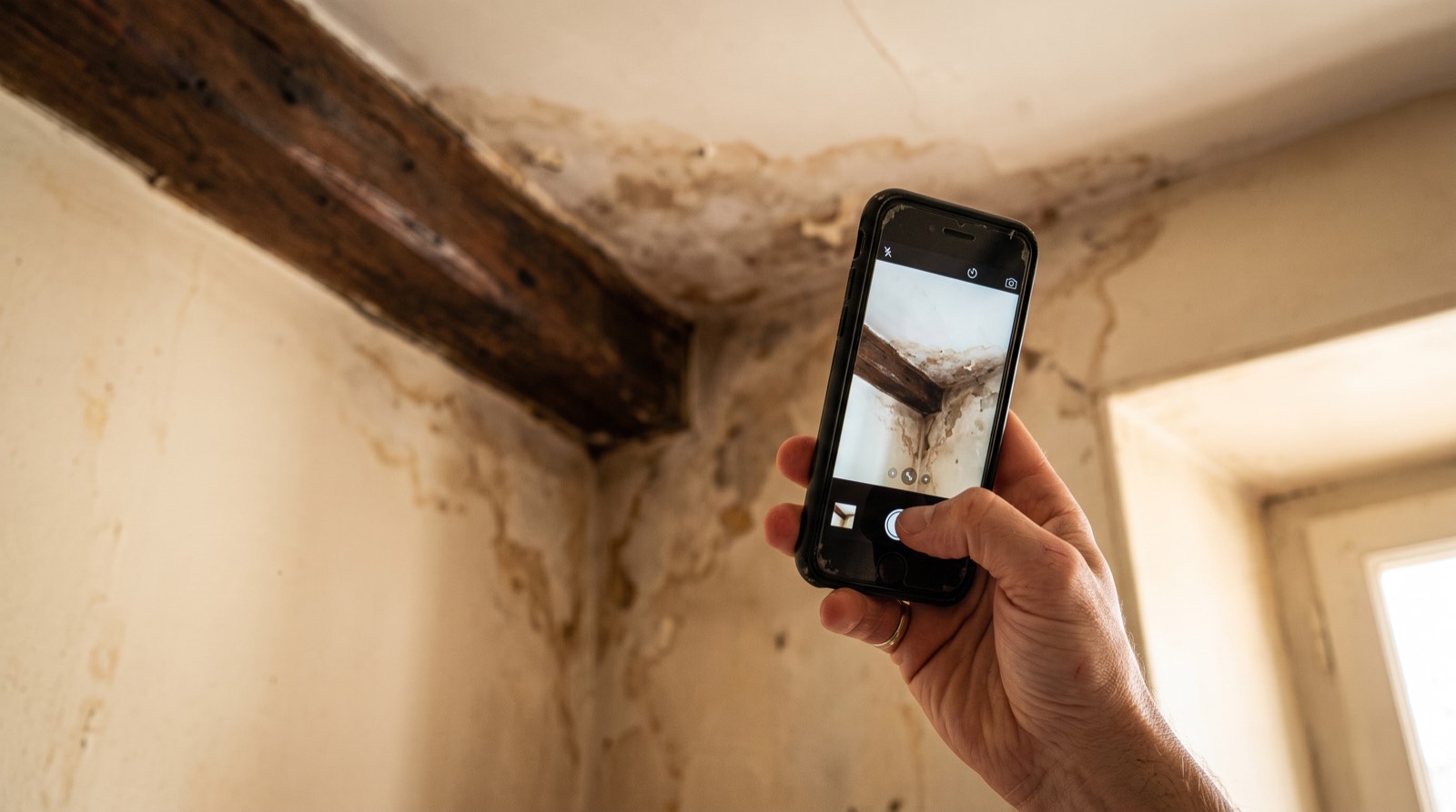 Main photographiant au smartphone une tache d'humidité au plafond, sous une poutre en bois, pour documenter un sinistre avant déclaration